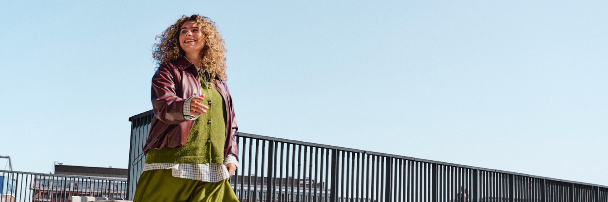  Model in a flowy green satin skirt, green sweater and burgundy jacket walking down a staircase
